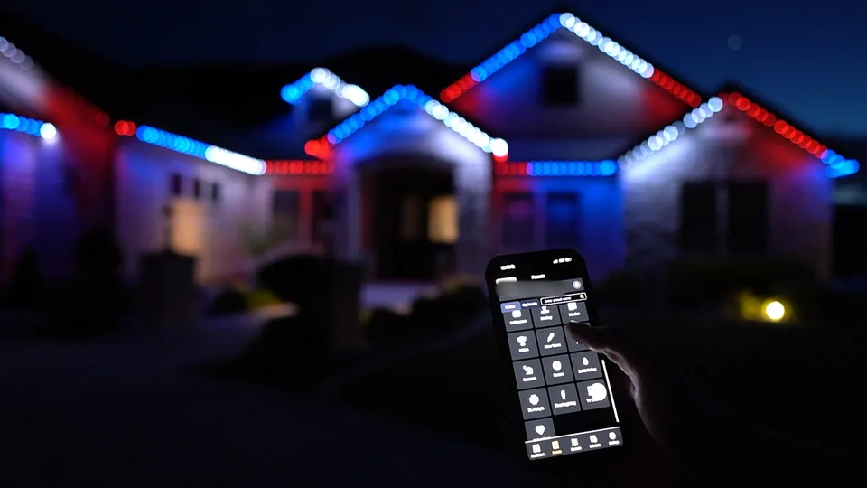 A hand holding up a phone with a grid of saved scenes on its screen, in front of a house at night whose roofline is lit in red, white and blue.