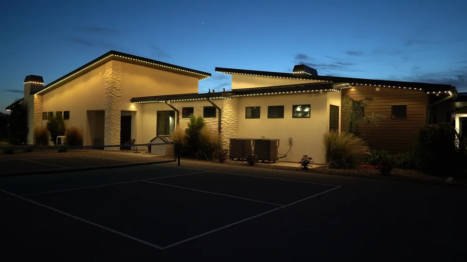 Warm white on a modern house, seen across a dark sport court at dusk.
