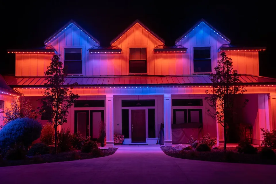 A white farmhouse at night washed in red, orange and pink, with blue and violet tracing its two outer dormer gables.