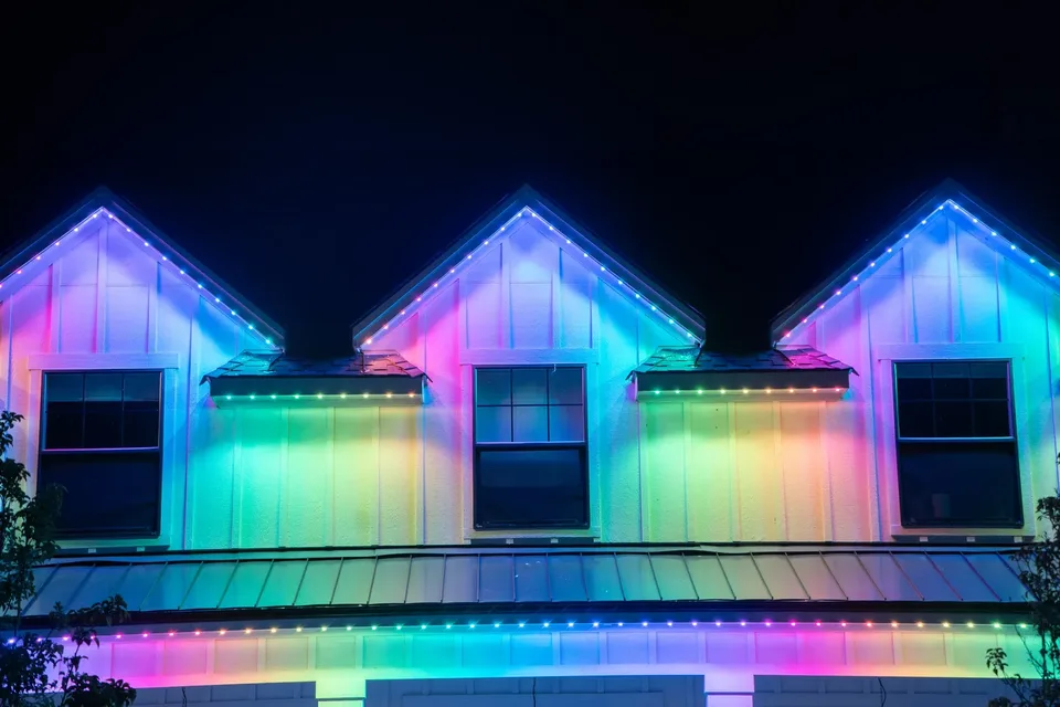 Three dormers above a metal porch roof at night, each peak outlined in light and the siding washed in a rainbow of pink, teal and gold.