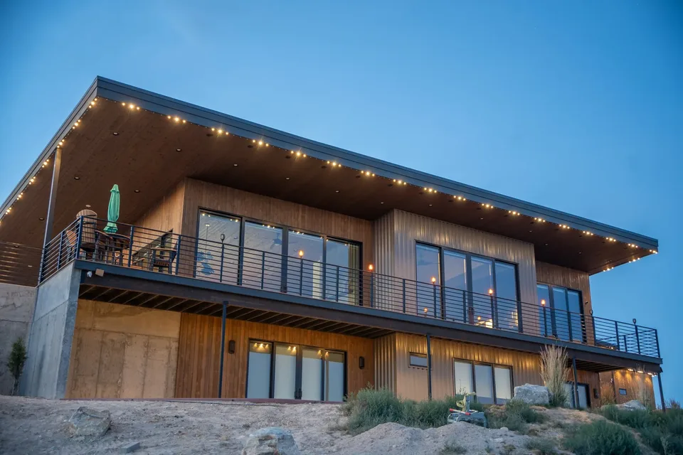Warm points along the underside of a long shed roof, over the deck of a modern wood-clad house at blue hour.