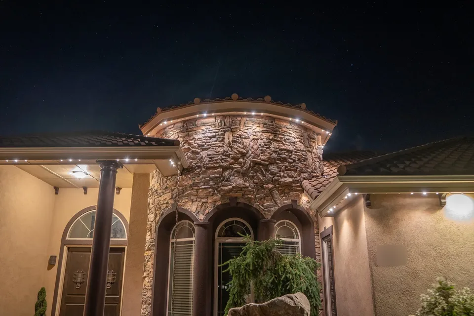 A curved stone entry tower at night under a tile roof, warm white lights following the curve of its eave and the eaves of the porch beside it.