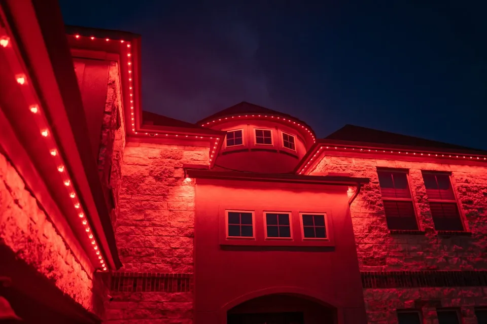 A stone house at night lit entirely in red, the light following a curved turret eave, the fascia and the stone walls below.