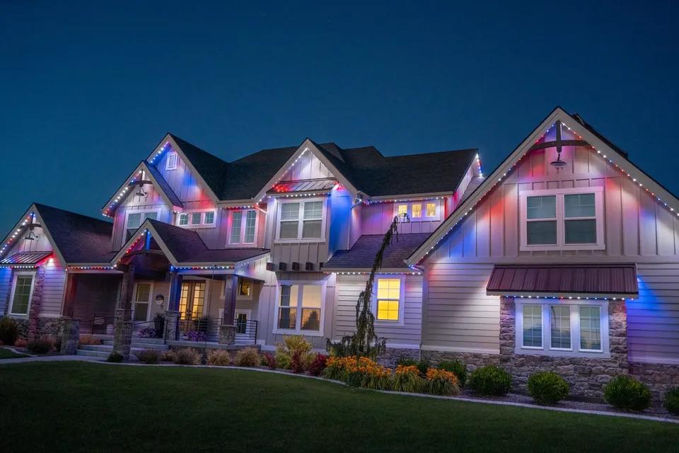 A tall two story farmhouse at dusk, red, white and blue light tracing every gable up to the highest peak.