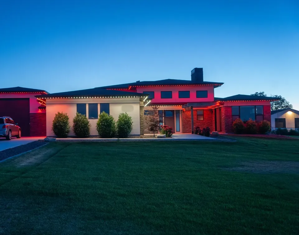 A modern two-story house at dusk with red lighting its upper walls and the brick wing at right, cream at left, over a wide lawn.