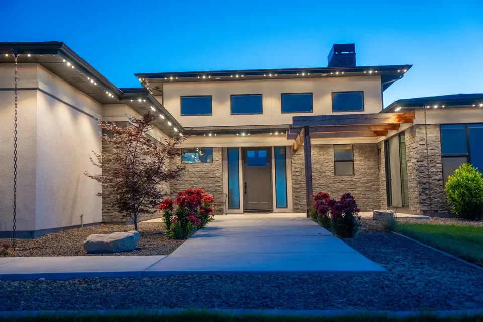 A two story stucco and stone house at dusk, small points of light along the underside of its roof edges, a flat gravel bed and a concrete walk below.
