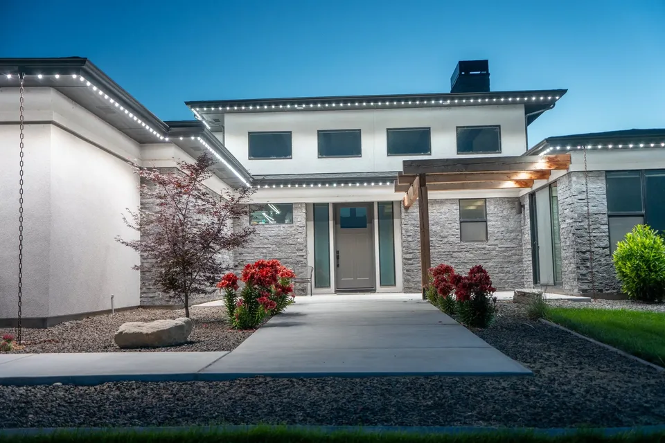 A low roofed stucco and stone house photographed head on down the entry walk at dusk, cool white light along every deep eave and the entry canopy, red flowers in the beds either side.