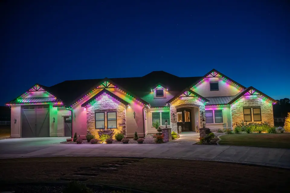 A wide single story house at night with each gable and eave lit a different pastel color, pink, green and violet, and the drive lit warm below.