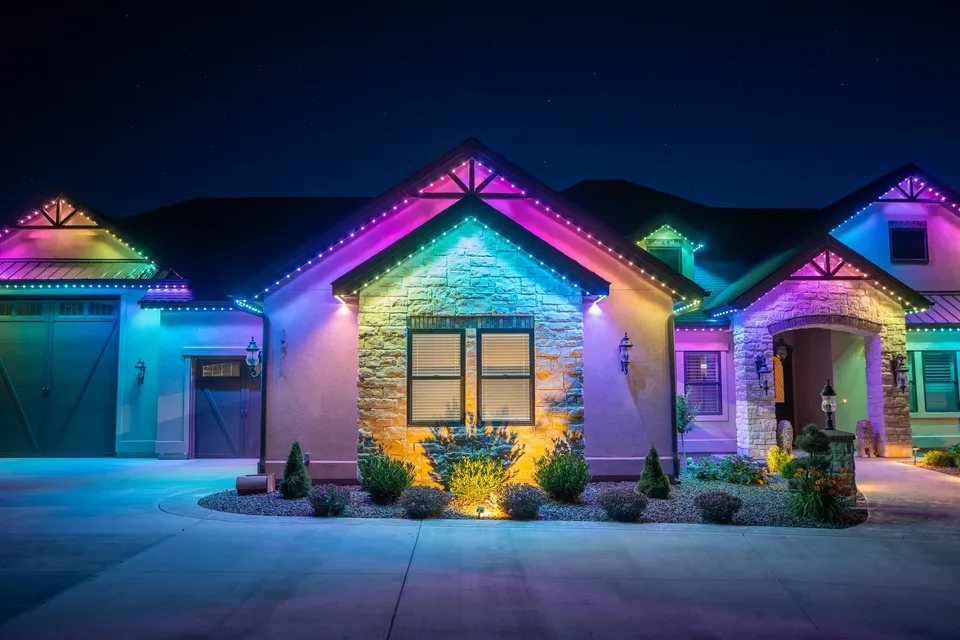 A stone and stucco home at night with pink, teal and violet light on every gable, the garage, the entry arch and the dormer.