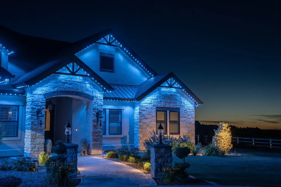 A stone and stucco house at night with its gables and timber trusses traced in cyan blue, a single tree uplit warm at the right and the last of the sunset on the horizon.