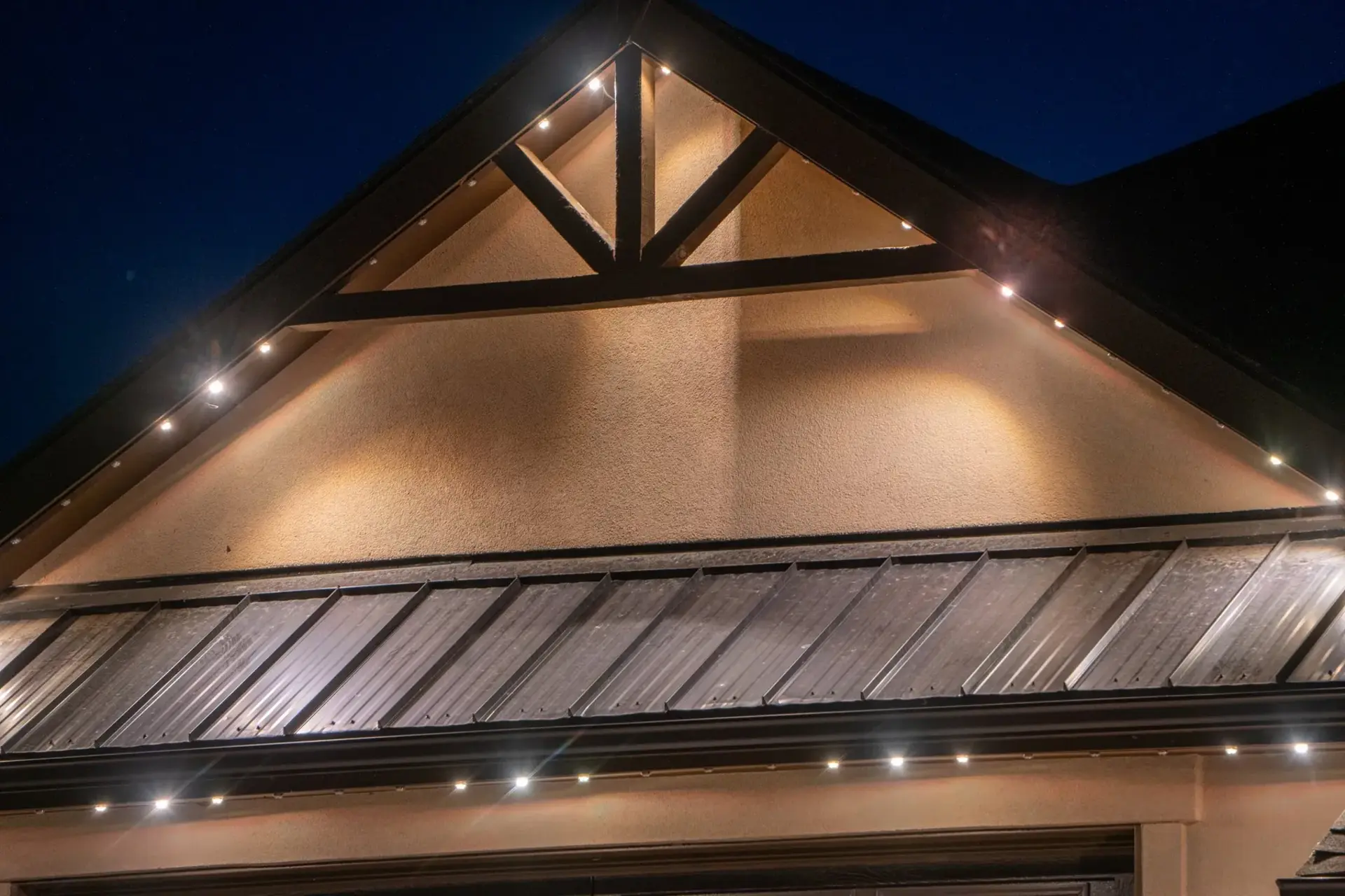 A close view of one gable at night. Small points of light sit in a channel under the roof edge and wash warm light down the stucco face below.