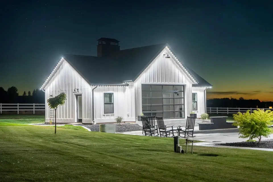 A board and batten farmhouse at dusk, its two gables and the eave between them traced in cool white light, with a sunset on the horizon behind it.