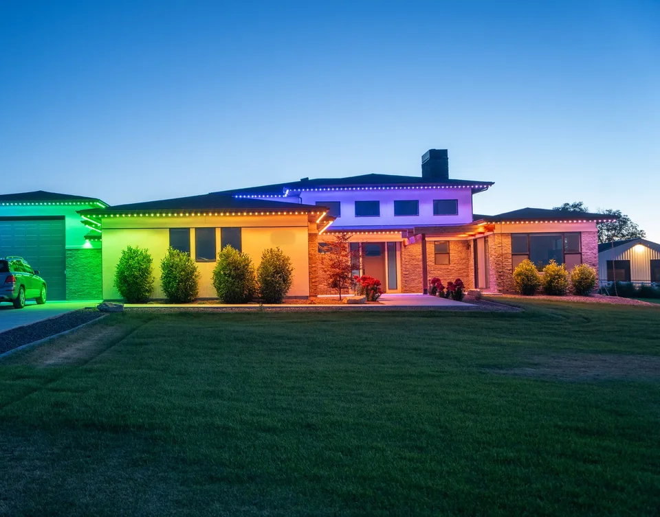 A long single story house at blue hour with its roofline running a continuous spectrum, green at the garage end through yellow and amber at the entry to red at the far wing, and blue along the upper roof.