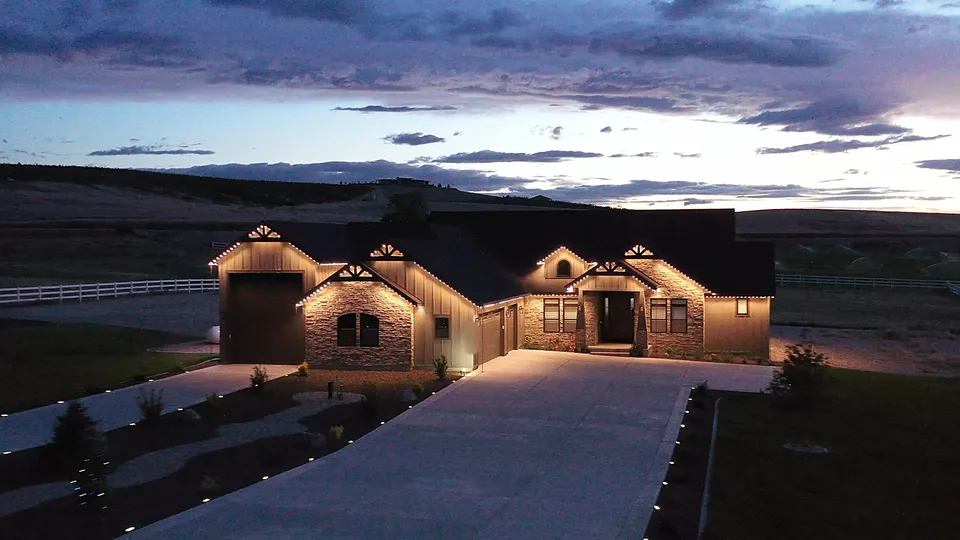 A long ranch front lit warm white along every eave and gable, under a purple and orange sunset on the ridge behind it.