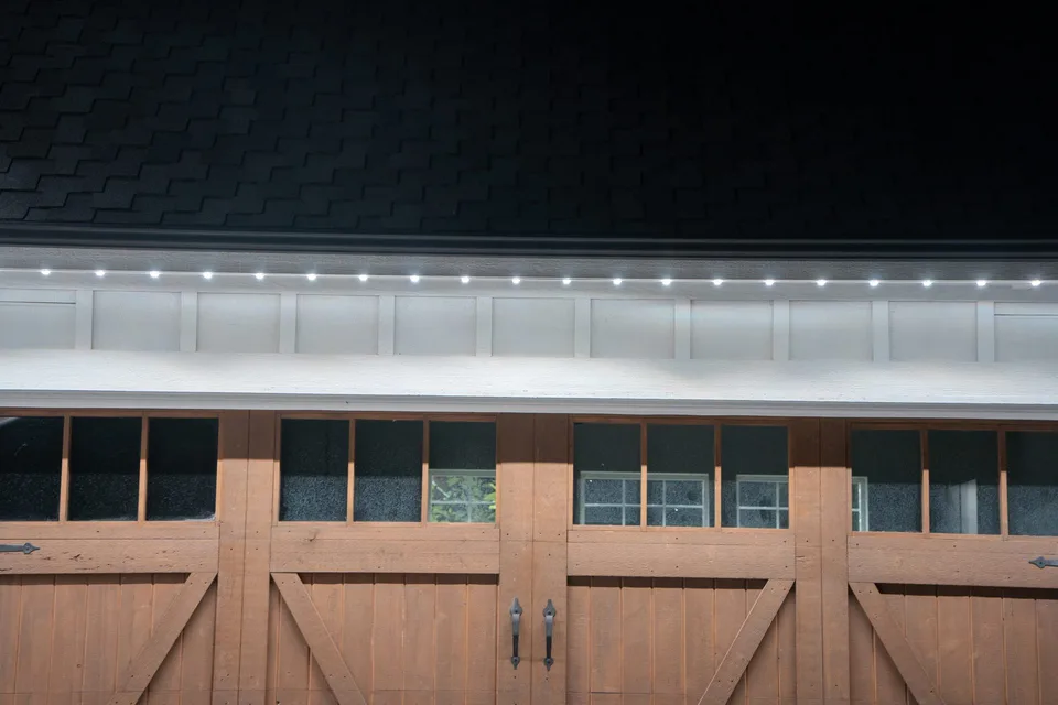 A cool white run along a garage eave above wood carriage doors, throwing a soft wash down the wall.