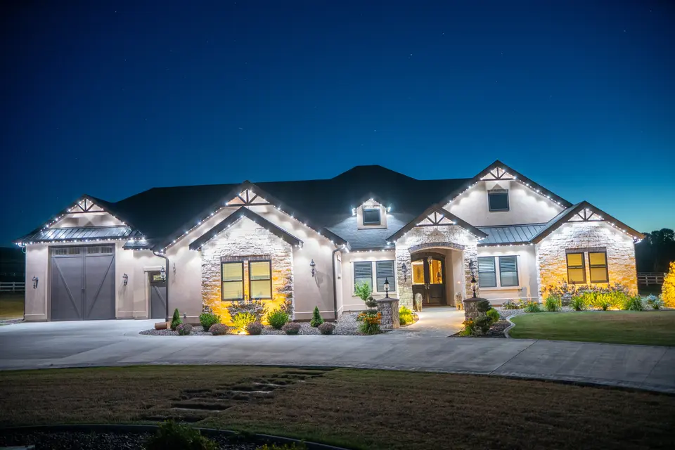 A wide single story house at twilight, cool white light running along every gable, the dormer, the porch roof and the garage, with the beds below it uplit.