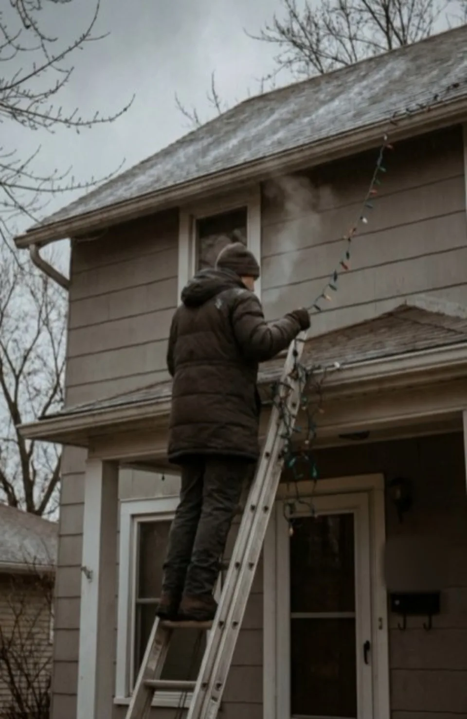 A man on an extension ladder in winter, clipping a string of lights along the edge of a roof.