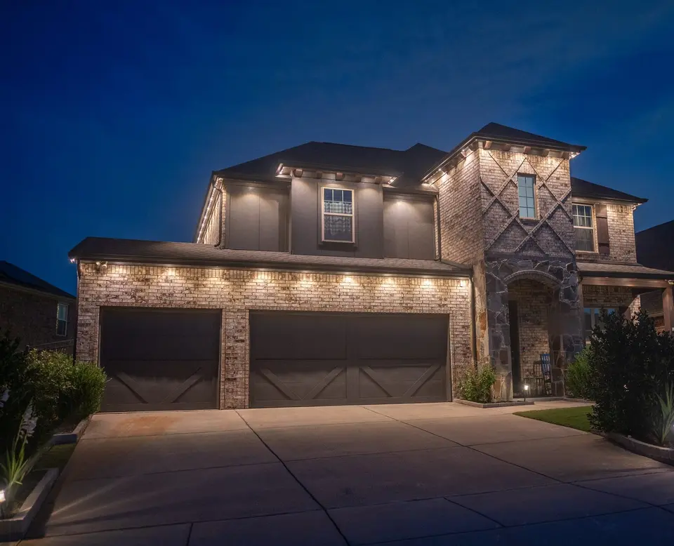 A two story house at dusk with warm light under the eave above the garage doors, along the upper roofline, and around an entry tower at the right.