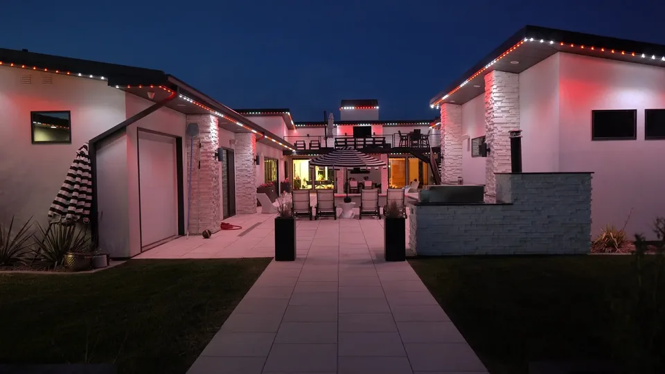 A white courtyard house at night, red and white points along its roof edges and a red wash across the walls, a pale tile path running back to a seating area.