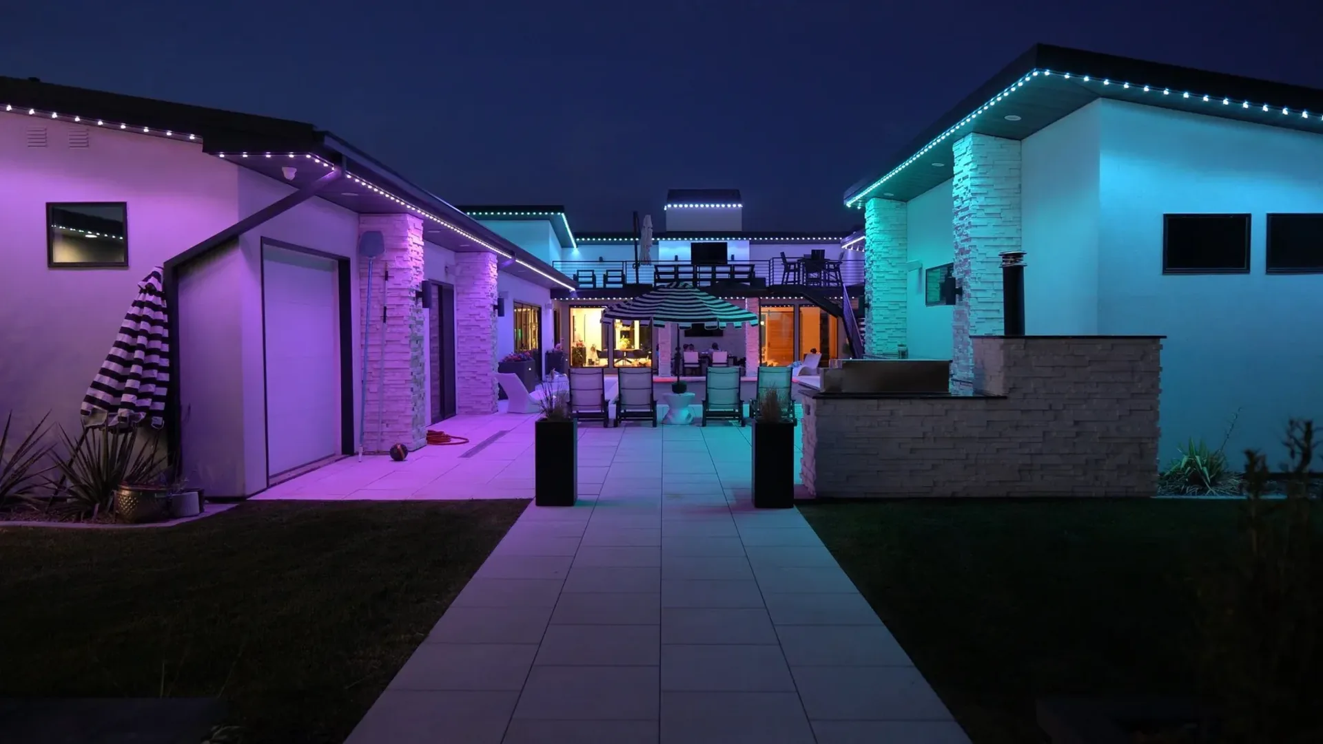 A lit walkway at night between two wings of one property, the left wing washed magenta and the right one teal, the roof edges of both picked out in the same colors.