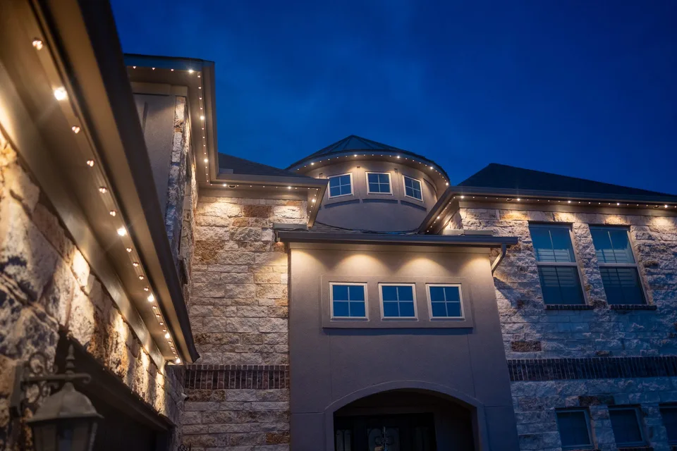 A stone estate front at blue hour with warm light under every eave, carried around a turret roof and out along the wings on both sides.