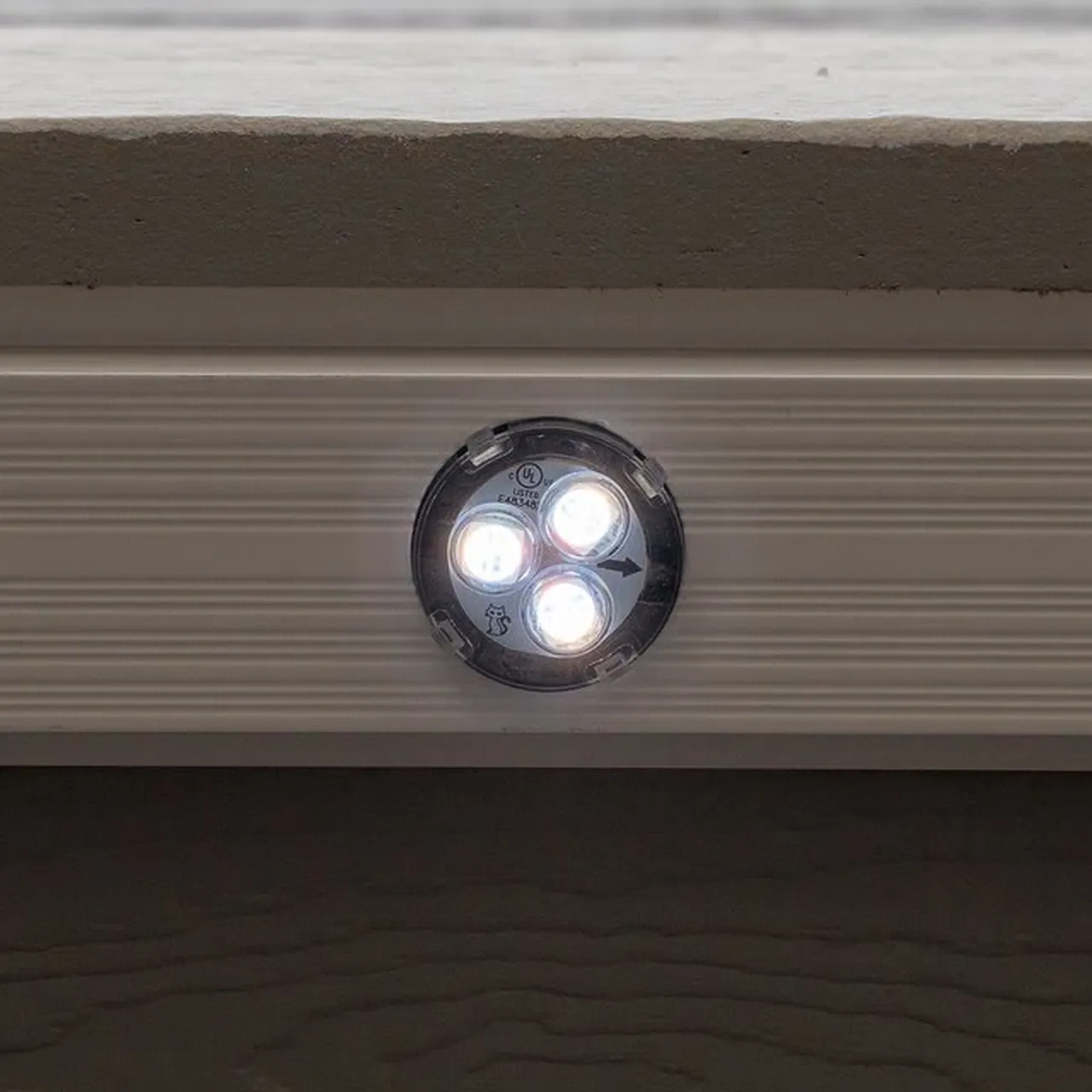One light switched on inside a white ribbed channel under a fascia board: a round clear lens over three glowing LEDs, sitting flush in the channel.